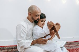 A father and son in traditional attire share a moment of prayer with the Quran indoors.